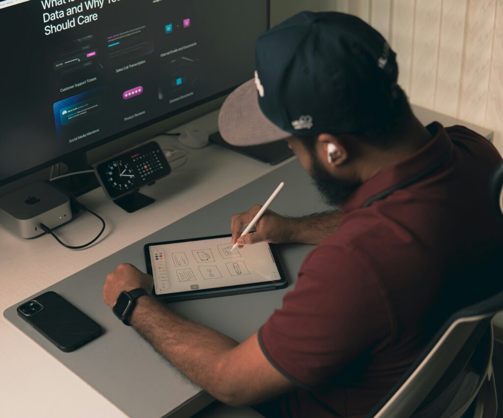 Designer esboça telas de aplicativo com caneta digital em um tablet, ao lado de monitor, celular e computador sobre a mesa